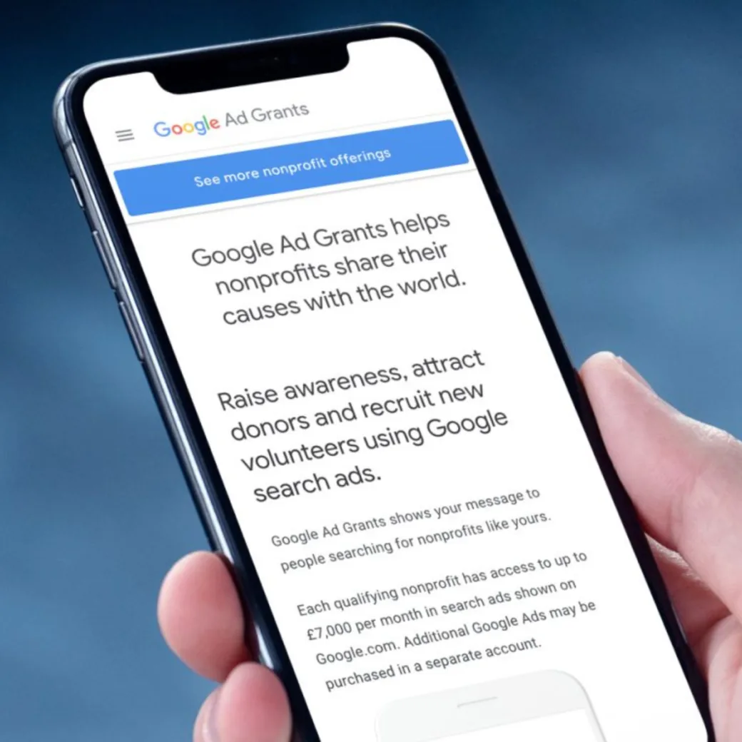 Hand holding a smartphone displaying a Google Ad Grants page. The screen explains how nonprofits can use Google search ads to raise awareness, attract donors, and recruit volunteers.