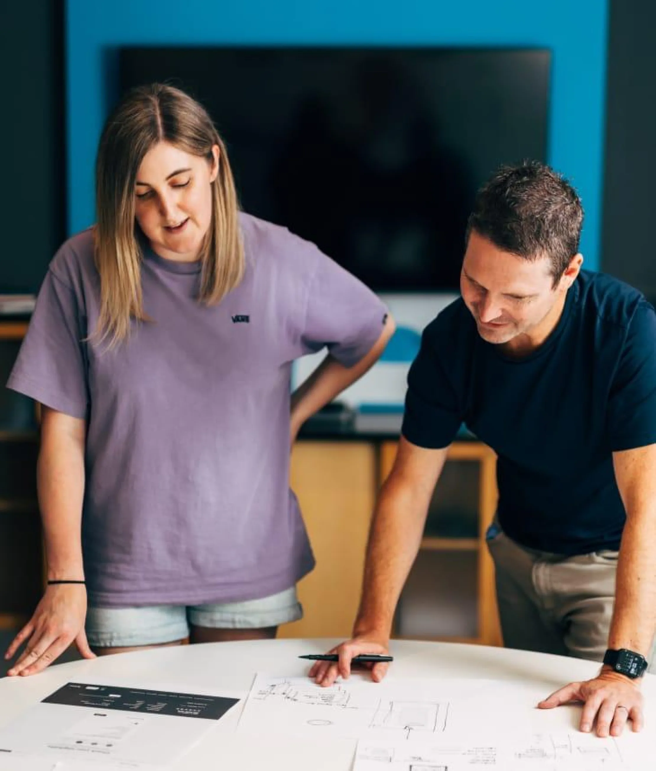 Two people standing at a table reviewing website wireframes and design plans, collaborating in a modern office setting.