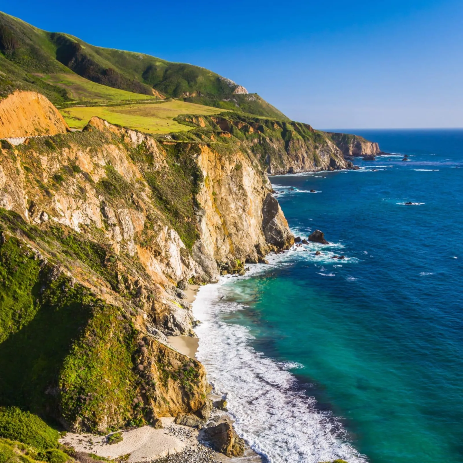 Sunlit coastal cliffs with green hills above turquoise ocean waves along a rugged shoreline under a clear blue sky.