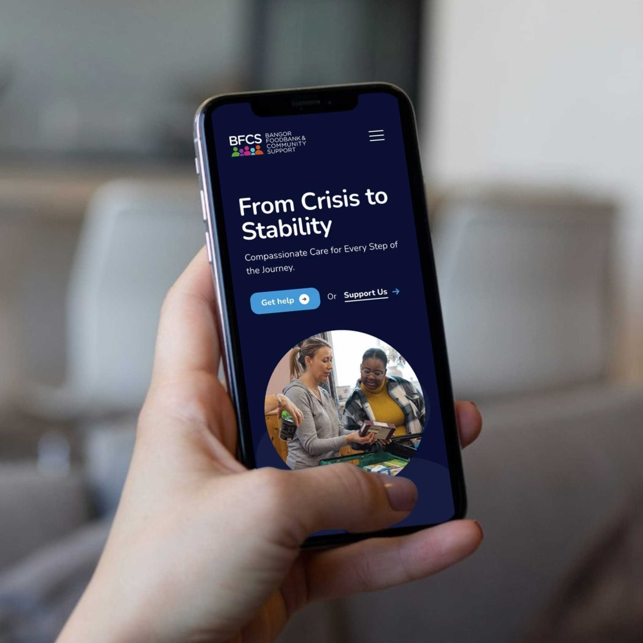 Hand holding smartphone displaying Bangor Foodbank website with headline 'From Crisis to Stability' and an image of two people talking.