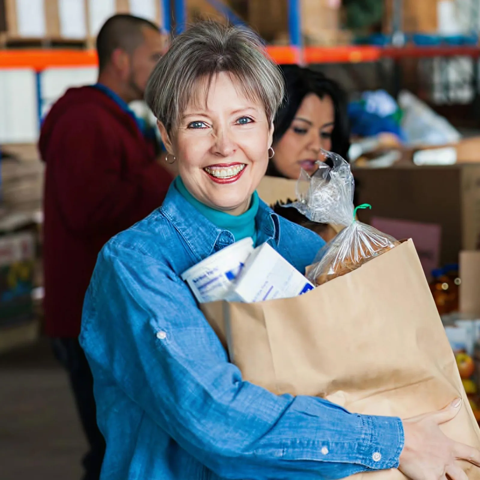 Smiling woman holding a paper bag of groceries in a foodbank setting with volunteers working in the background.